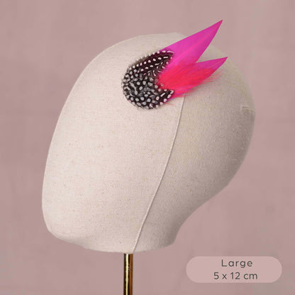 A decorative hair accessory with two bright pink feathers and a black-and-white spotted feathers, displayed on a beige fabric mannequin head against a dusty pink background. A text label in the bottom right reads 'Large 5 x 12 cm'.