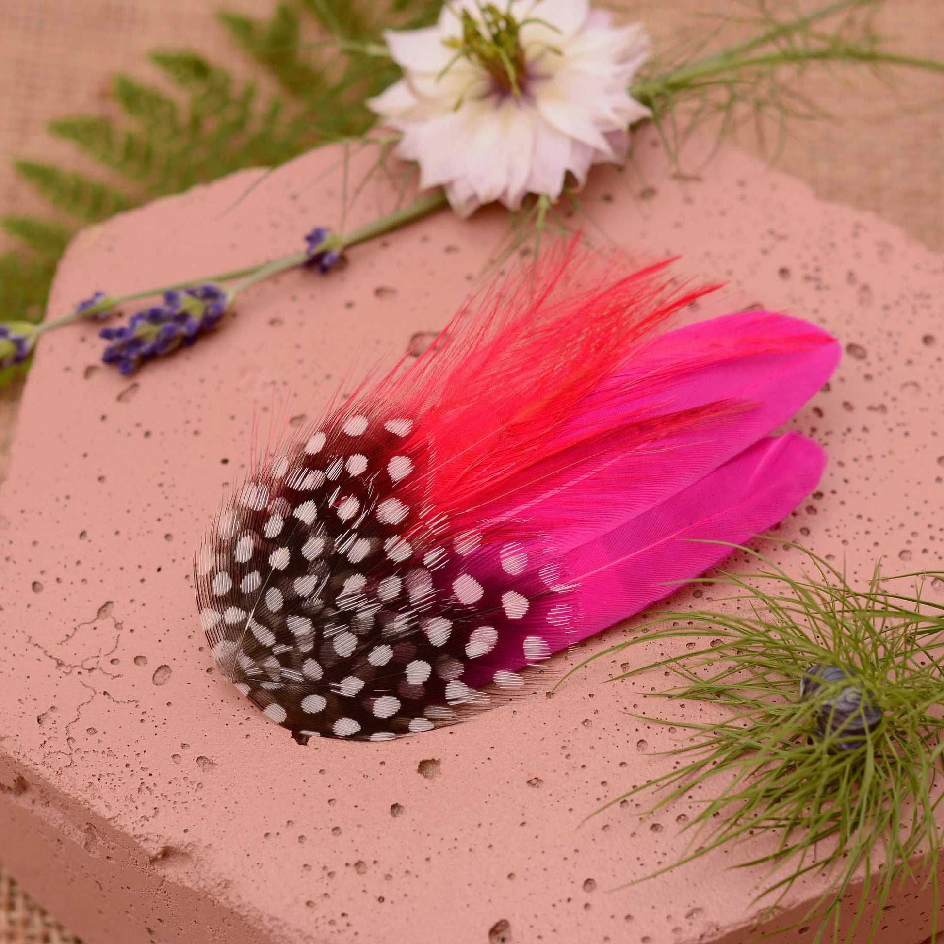 A decorative feather hair clip in bright hot pink feathers and black feathers with white spots, styled on a soft pink stone with flowers and greenery.