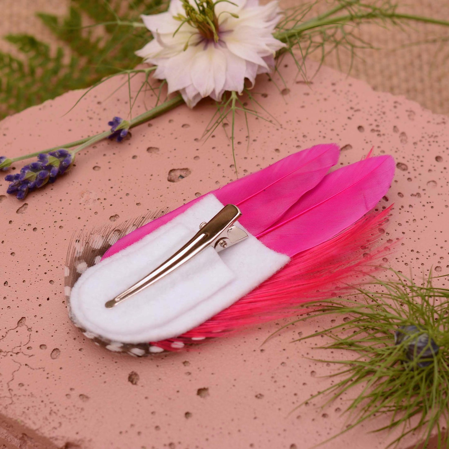 A decorative feather hair clip in bright hot pink feathers and black feathers with white spots. Displayed upside down to show the white felt base and alligator style clip. Styled on a soft pink stone with flowers and greenery.