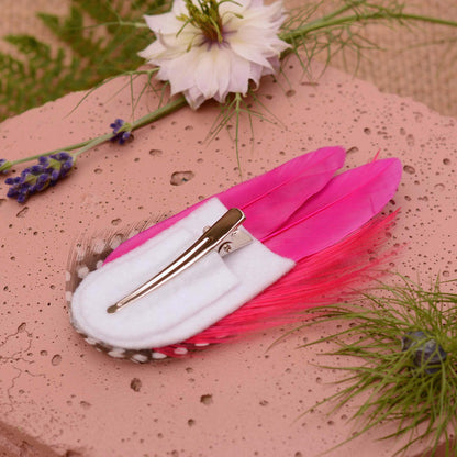 A decorative feather hair clip in bright hot pink feathers and black feathers with white spots. Displayed upside down to show the white felt base and alligator style clip. Styled on a soft pink stone with flowers and greenery.