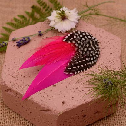 A decorative feather hair clip in bright hot pink feathers and black feathers with white spots, styled on a soft pink stone with flowers and greenery.
