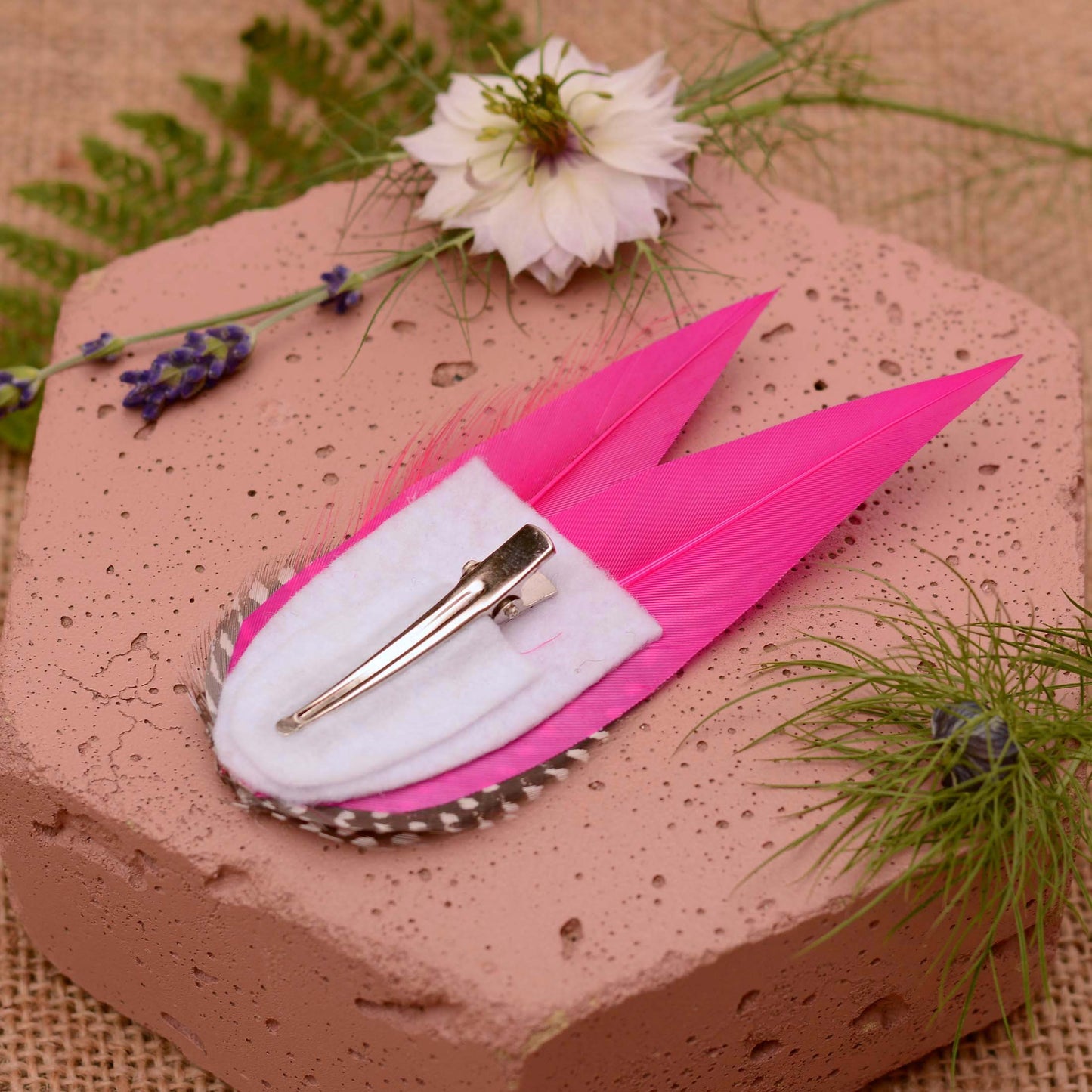 A decorative feather hair clip in bright hot pink feathers and black feathers with white spots. Displayed upside down to show the white felt base and alligator style clip. Styled on a soft pink stone with flowers and greenery.
