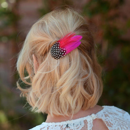 A blonde woman with a short wavy bob styled in a half-up do shown from the back, wearing a hot pink and black and white polka dot feather hair clip accessory.