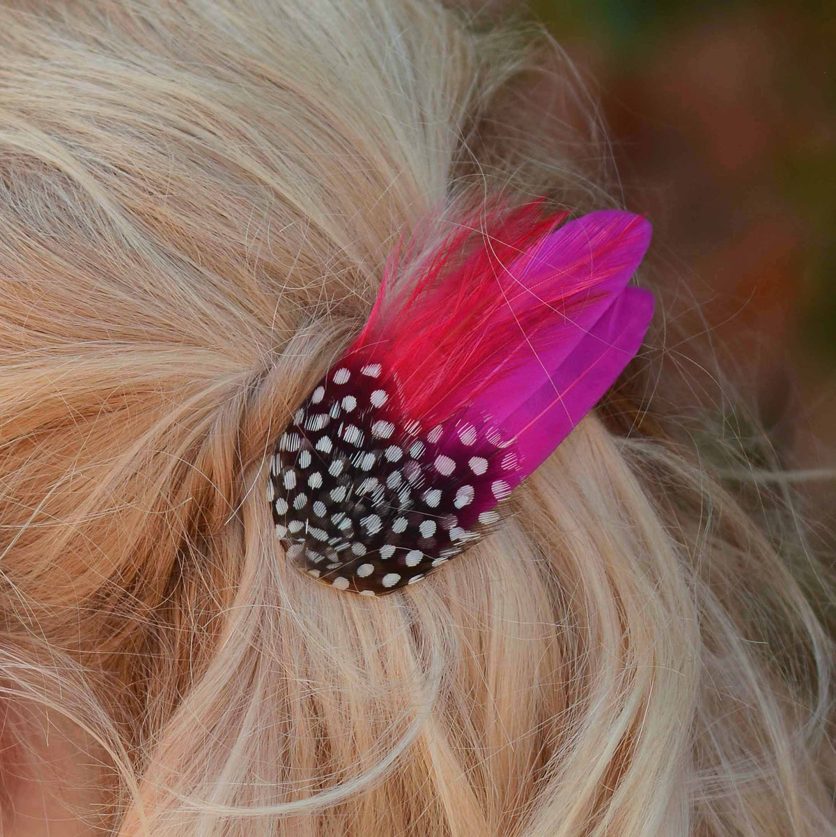 A close up of a blonde woman with a short wavy bob styled in a half-up do shown from the back, wearing a hot pink and black and white polka dot feather hair clip accessory.