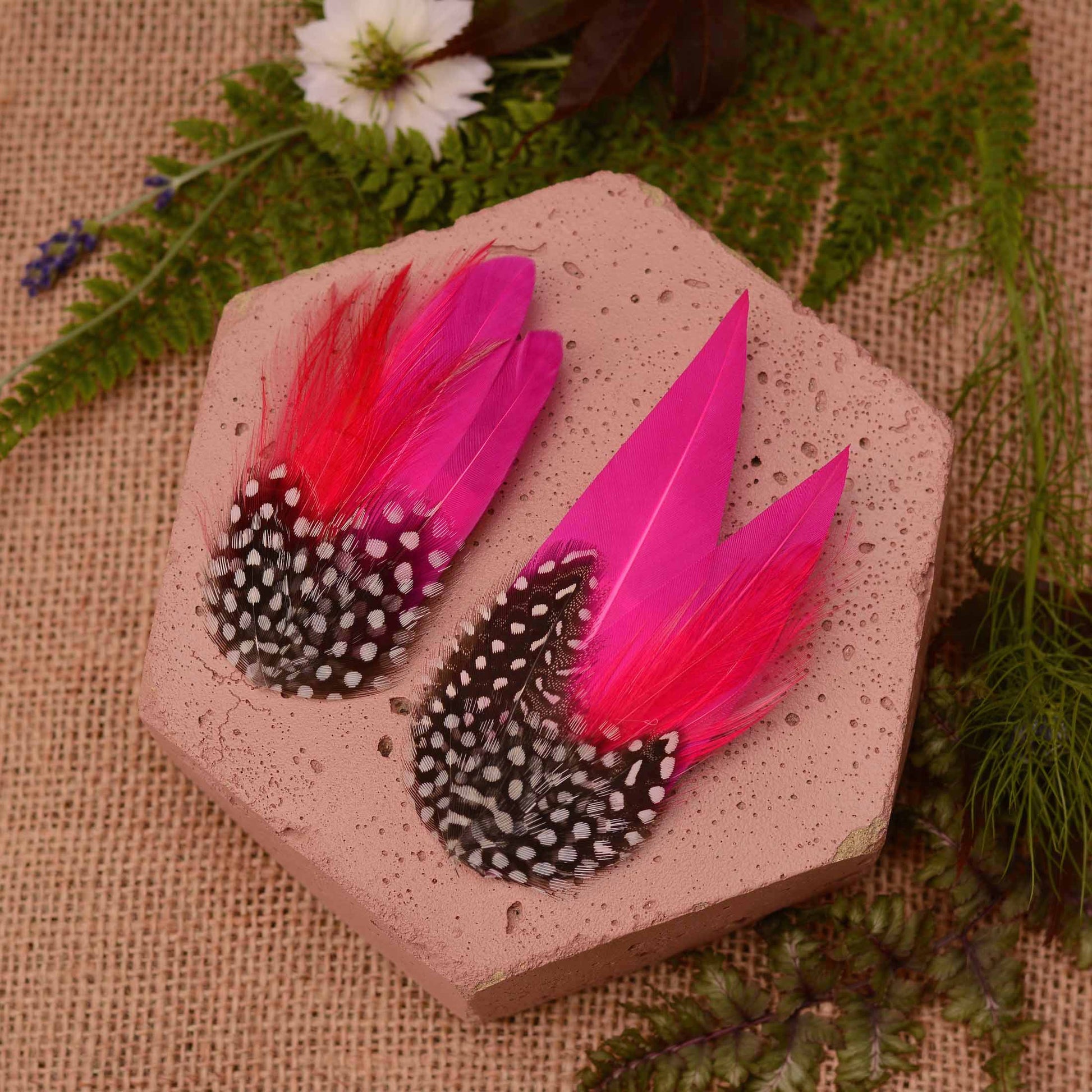 Two decorative feather accessories with bright pink feathers and black-and-white spotted feathers, displayed on a hexagonal stone slab with burlap and greenery in the background.