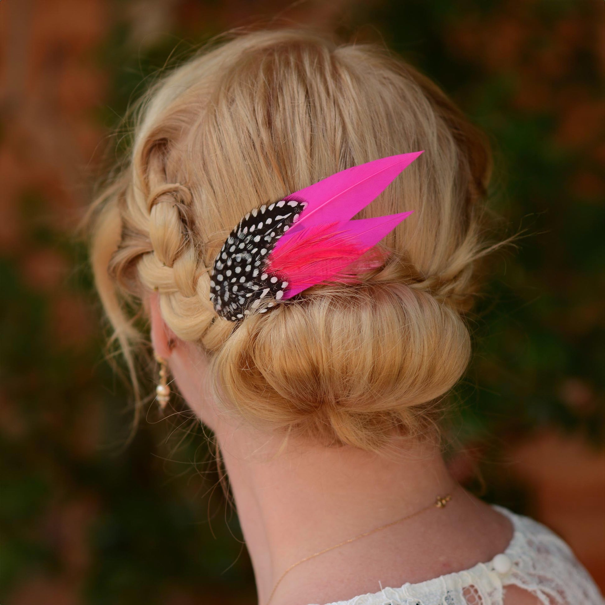 A blonde woman shown from the back wearing a braided half-up style bun with a bright pink feather hair accessory with black-and-white spotted feather accent.
