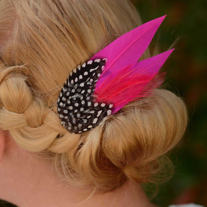 A close-up of a blonde woman's braided hair bun decorated with a black and white spotted feather clip and two bright pink feathers.