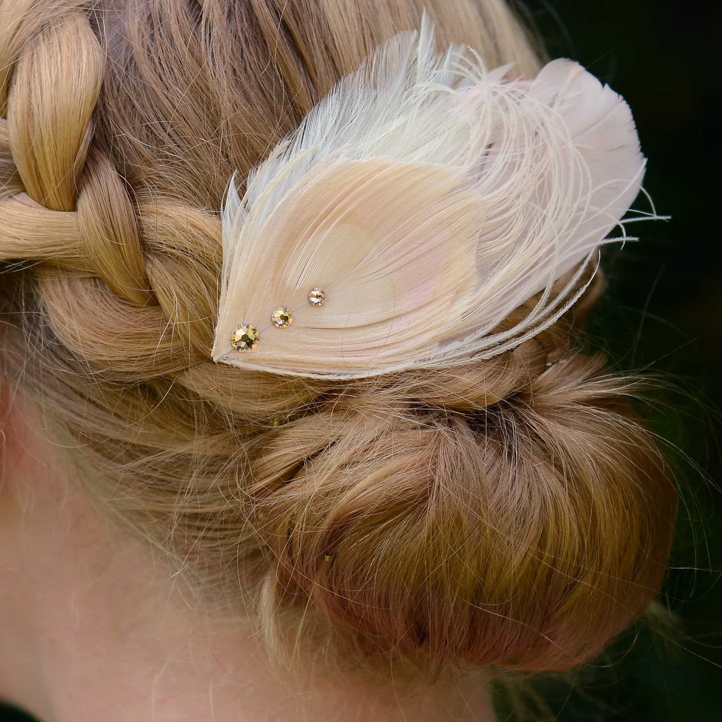 Close-up of braided hair with a decorative ivory and blush pink peacock Feather hair clip feather hairpiece  with crystals against a dark background