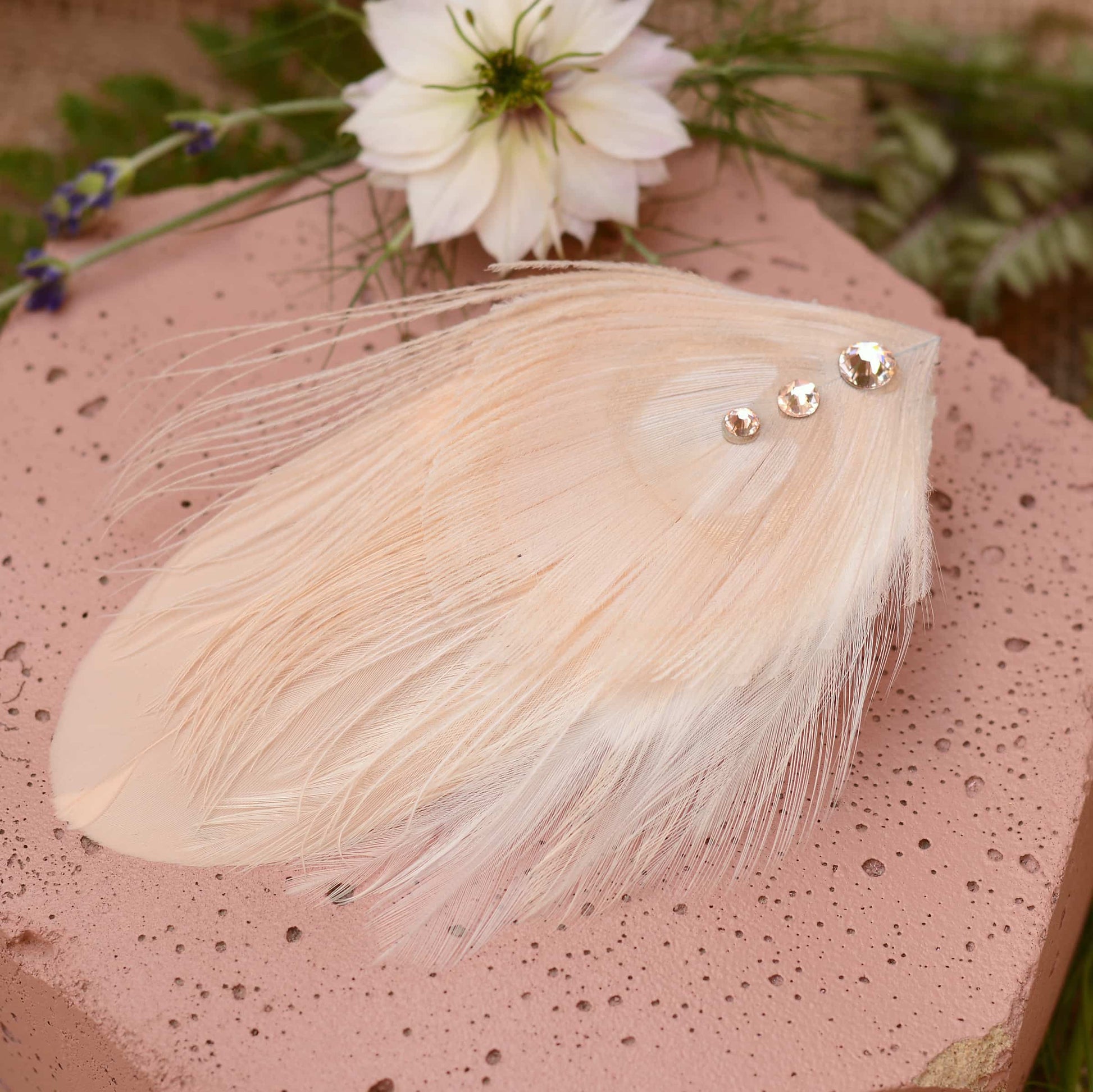 ivory and blush pink peacock Feather hair clip with crystals on a pink stone with flowers in the background