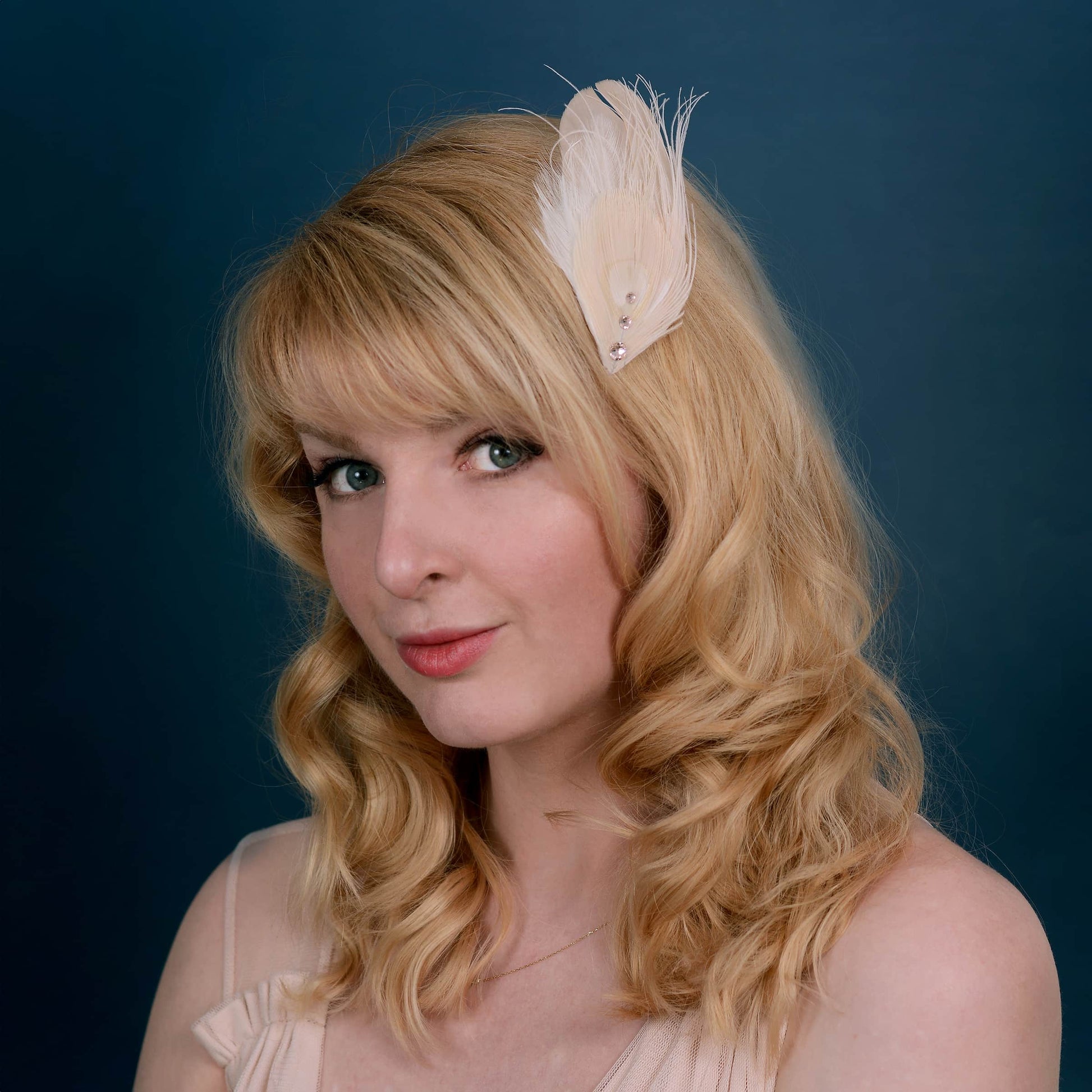 Woman with blonde curly hair wearing a ivory and blush pink peacock Feather hair clip with crystals clipped to the side of the head on a blue background