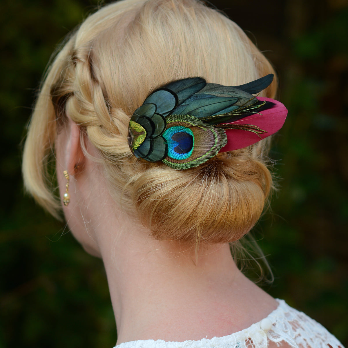 A blonde woman is shown from the back, wearing a decorative hair accessory with peacock feathers on a burgundy feather a blurred green outdoor background.