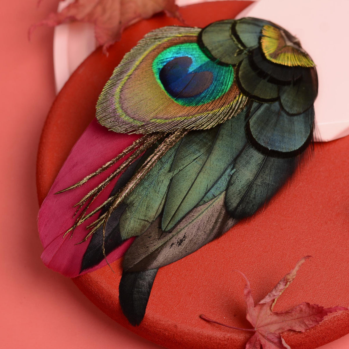 A close-up of a decorative feather accessory with iridescent green and blue peacock and rooster feathers layered over burgundy feathers, displayed on a brick-red surface against a coral pink background.