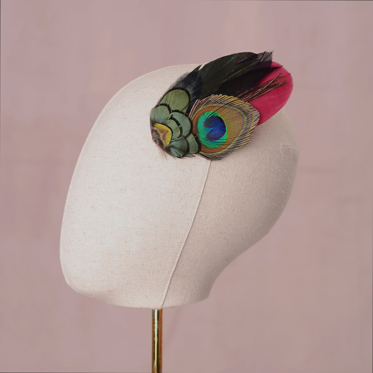 A decorative feather hair accessory with a burgundy feather and a large peacock-eye feather in green, blue, and black, styled on a canvas mannequin head.