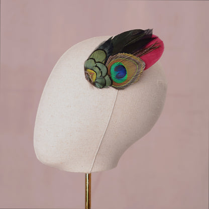 A decorative feather hair accessory with a burgundy feather and a large peacock-eye feather in green, blue, and black, styled on a canvas mannequin head.