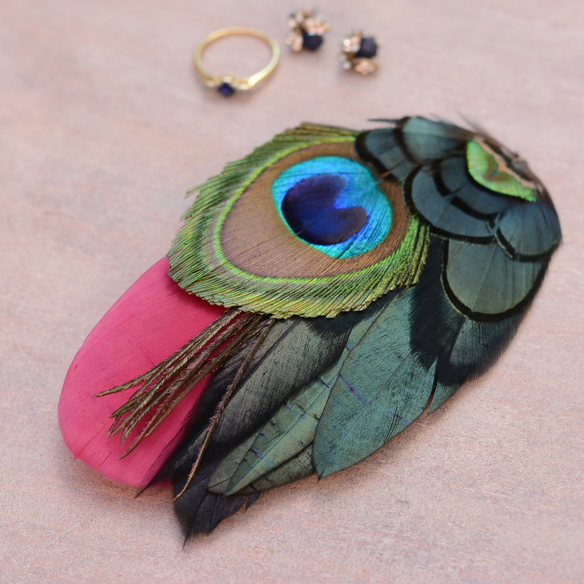 A decorative feather hair accessory with a burgundy feather and a large peacock-eye feather in green, blue, and black, styled on a beige surface with small gold jewellery pieces nearby.