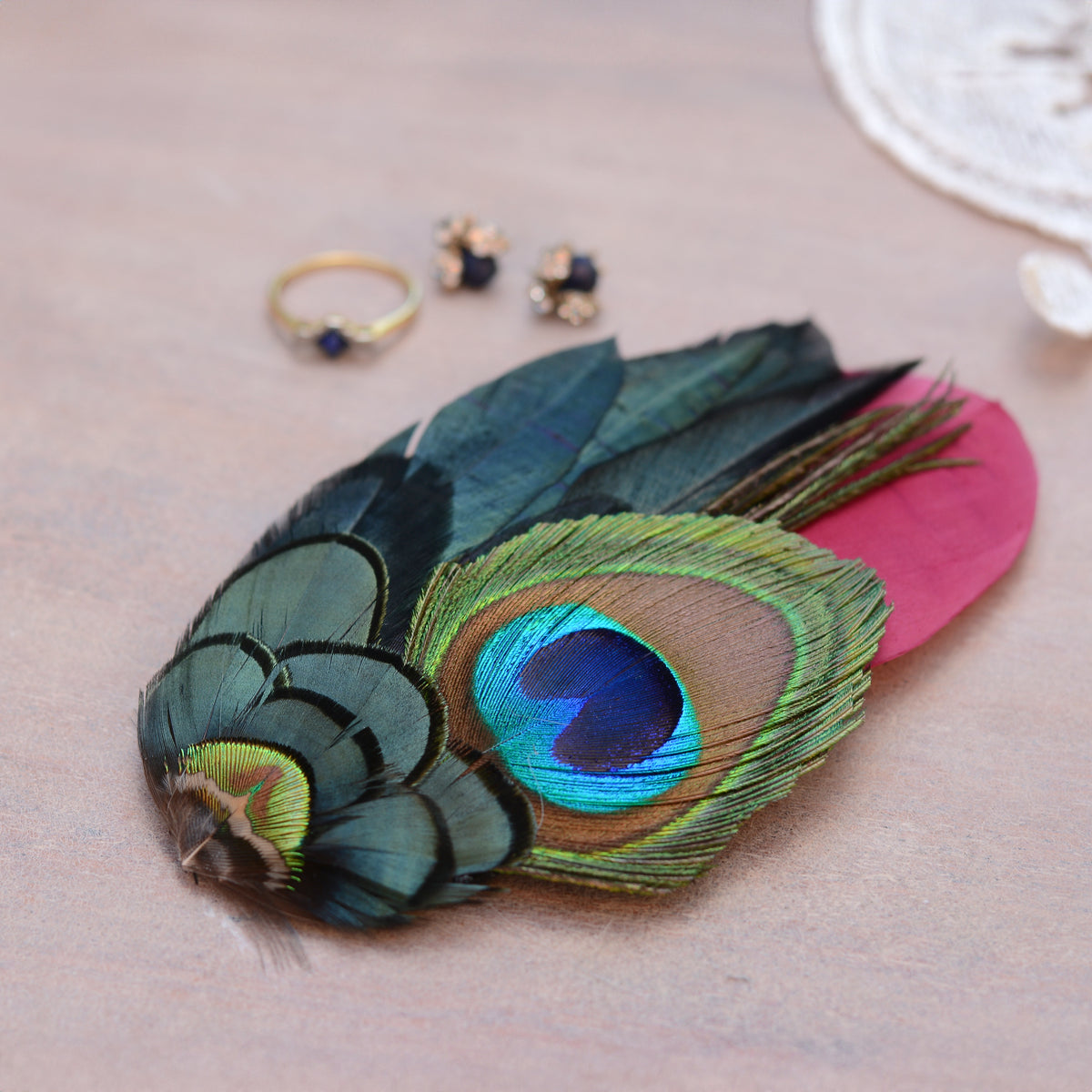 A decorative feather fascinator hair embellishment with a large peacock-eye pattern in blue, green, and gold, layered over a burgundy feather, displayed on a neutral beige surface with small jewellery props in the background.