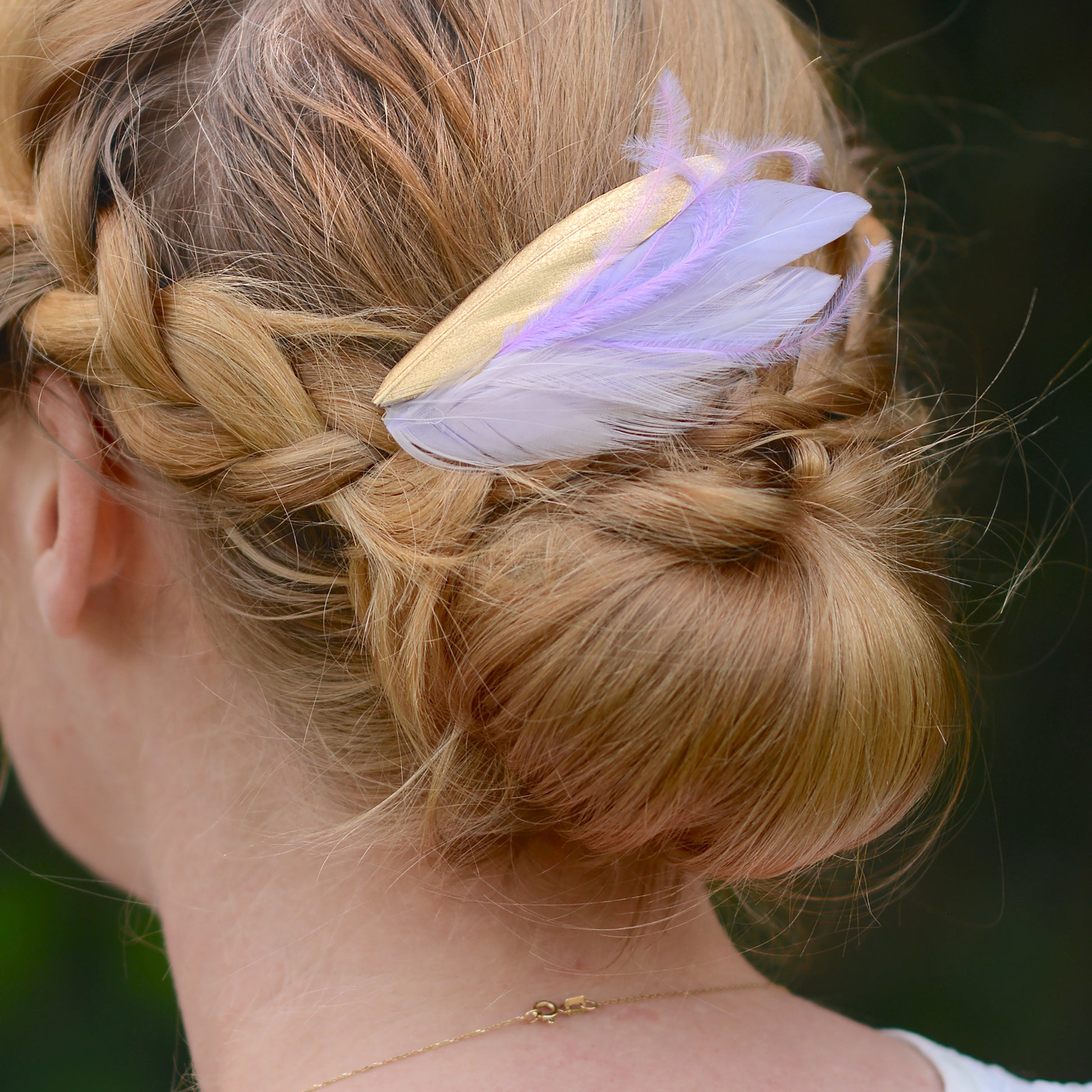 blonde braided ulow bun up-do viewed rom back decorated with a hair accessory of a small feather hair clip in pastel lavender purple and gold with different texture feathers