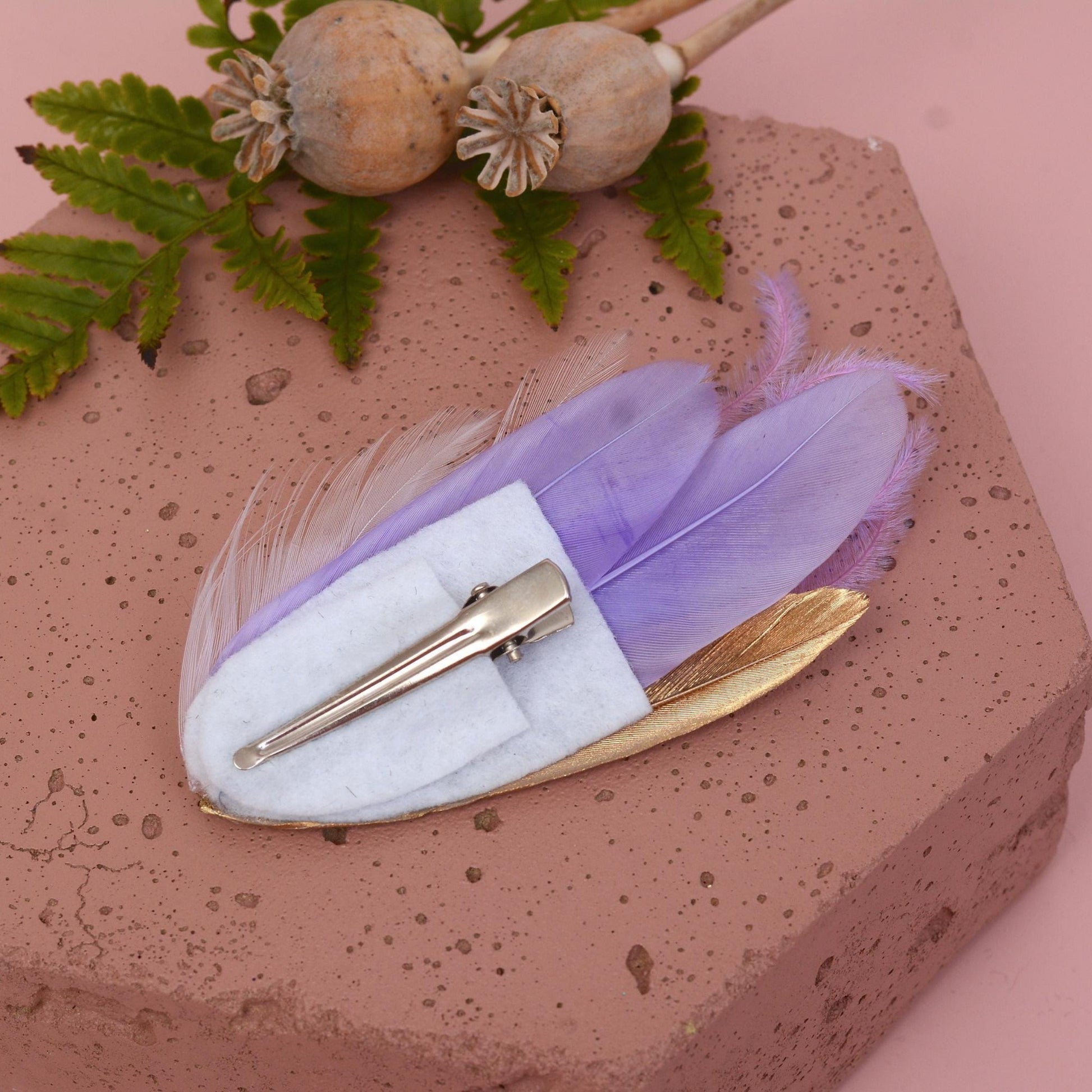 small feather hair clip in pastel lavender purple and gold upside down to show the clip attahcment  on a pink stone with flowers