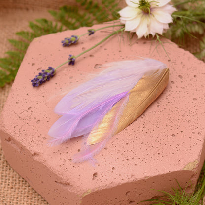 small feather hair clip in pastel lavender purple and gold with different texture feathers on a pink stone with flowers
