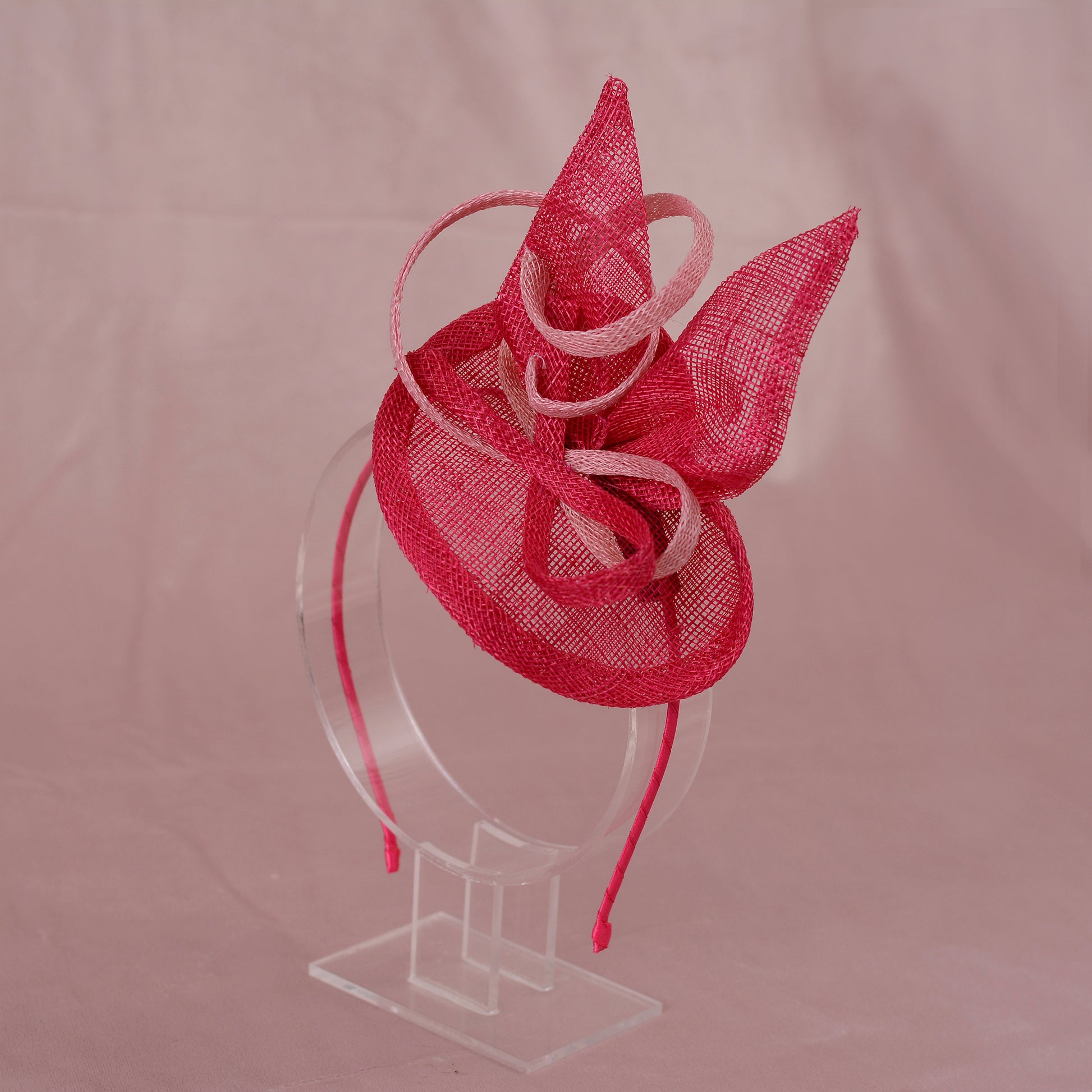 A pink sinamay side perching fascinator in two shades of tonal pink displayed on a clear acrylic stand against a soft pink background.