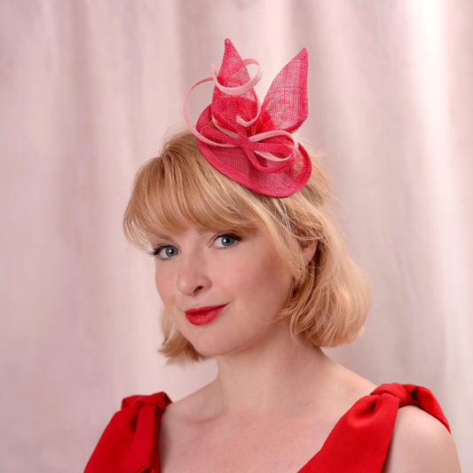 A woman with short blonde hair wearing a bright pink fascinator hat and a red dress, photographed against a soft pink draped background.
