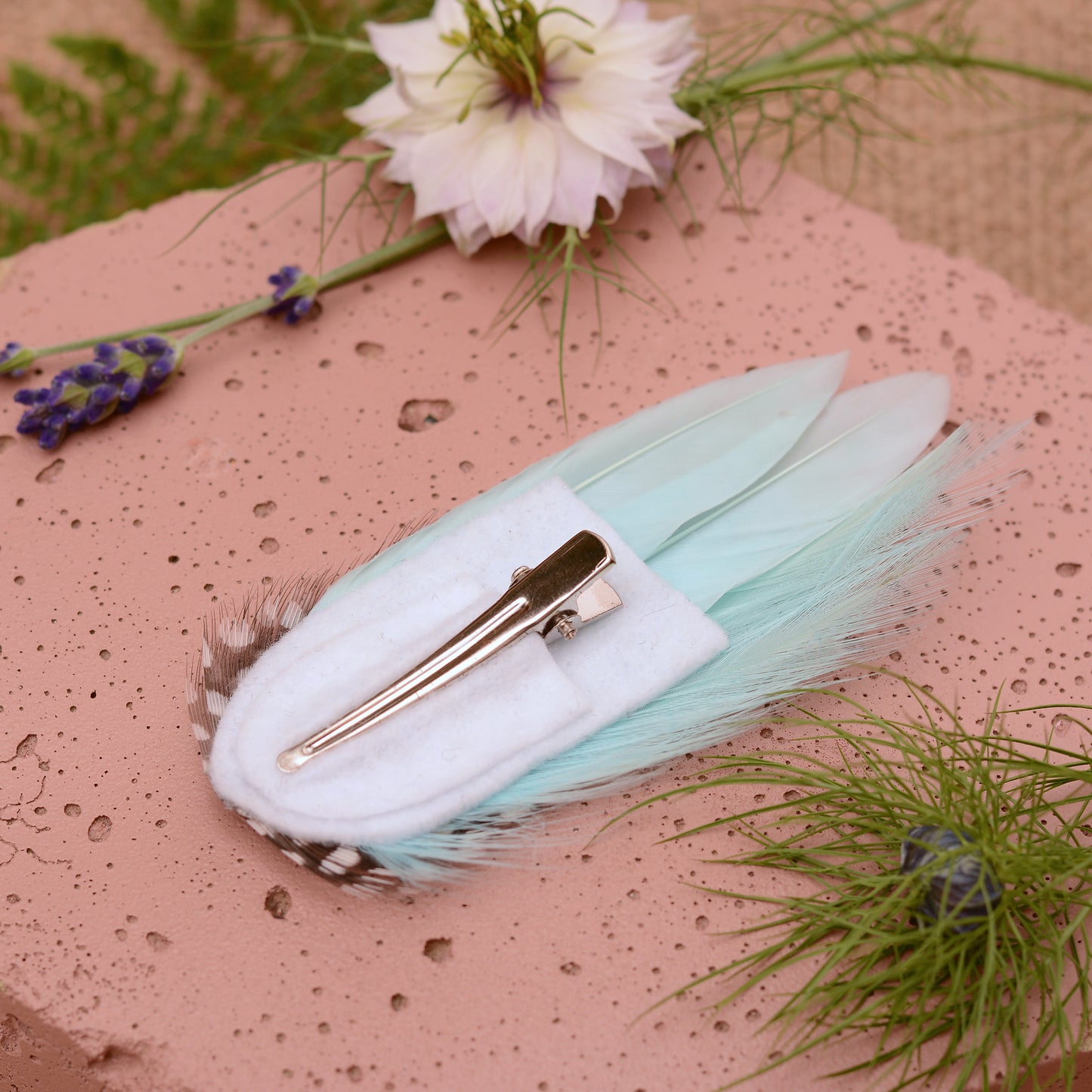 Mint green feather hair clip on a pink stone with flowers and plants around