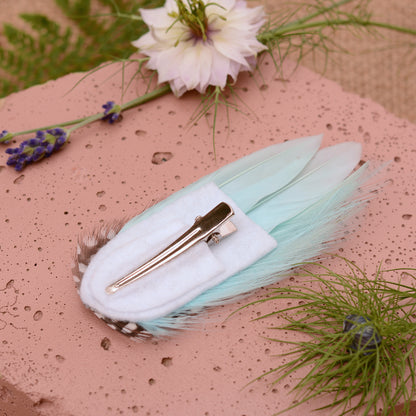 Mint green feather hair clip on a pink stone with flowers and plants around