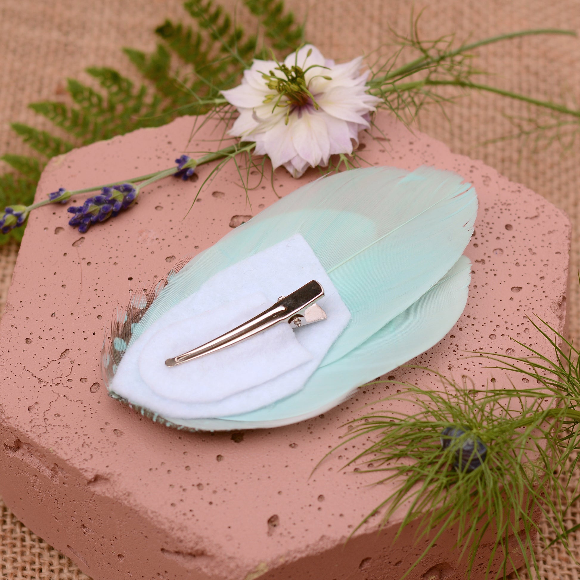 Mint green feather hair clip on a pink stone with flowers and plants around