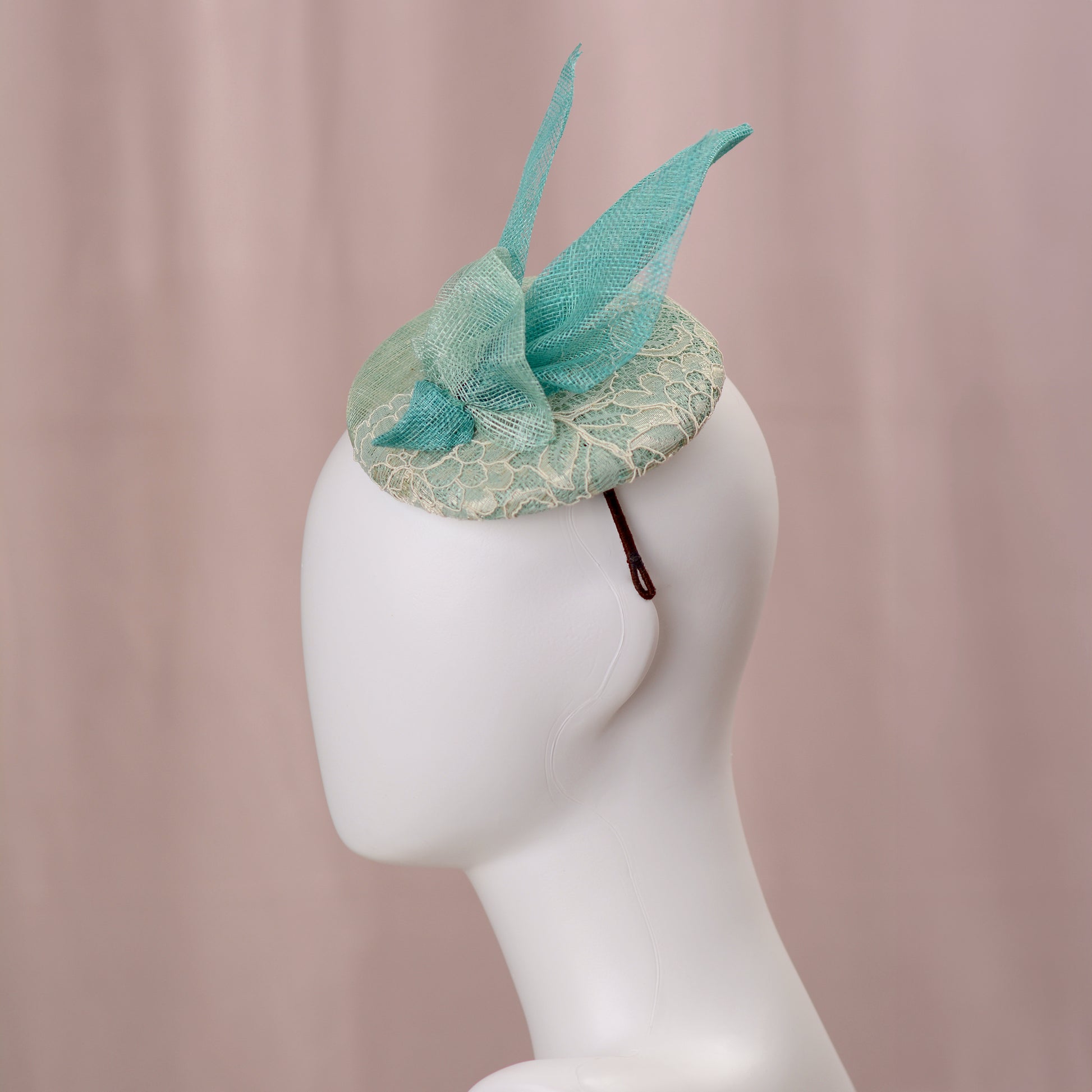 A mint green  and turquoise teal fascinator headpiece with sheer sinamay straw abstract bird wings and a lace trim, displayed on a white mannequin head against a soft pink background.