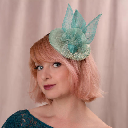 A woman with short pink hair wears a duck egg and teal fascinator hat with with lace and sinamay trim, photographed against a soft beige curtain background.