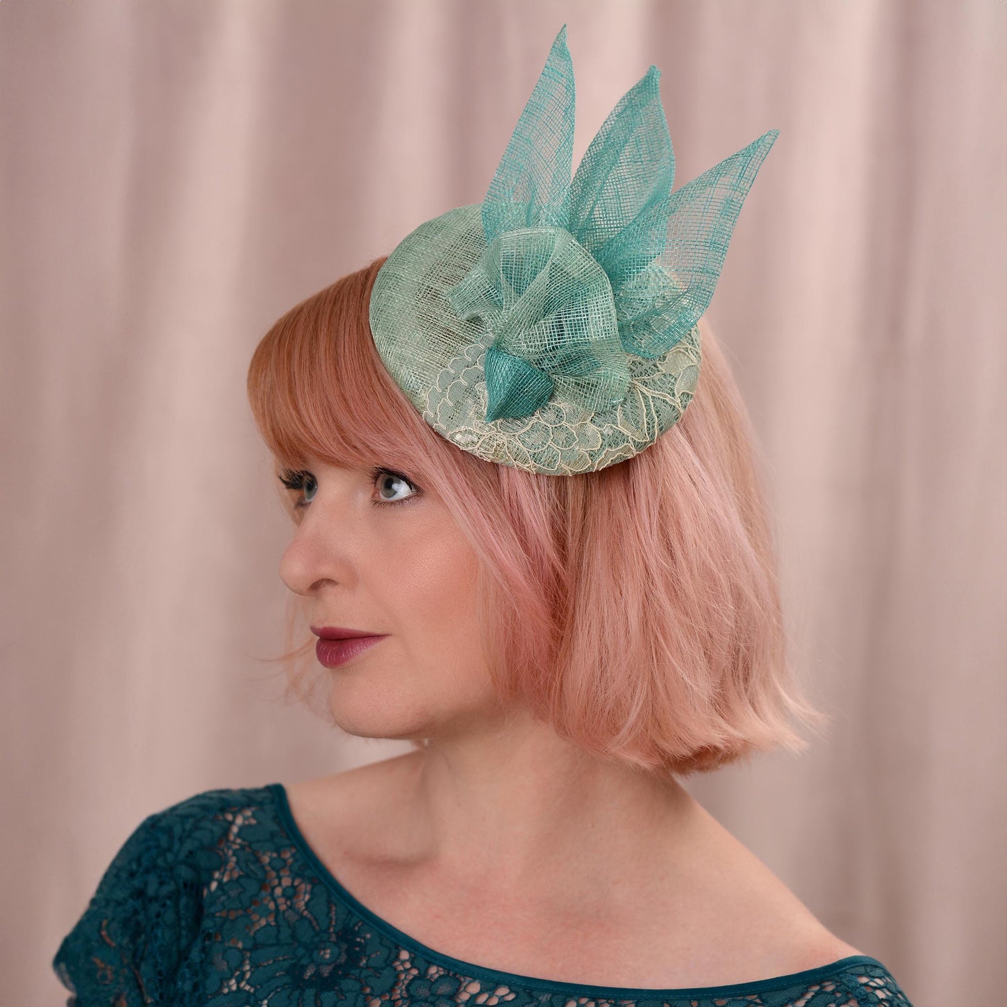 A woman with short pink hair wears a duck egg and teal wedding fascinator hat with with lace and sinamay trim, photographed against a soft beige curtain background.