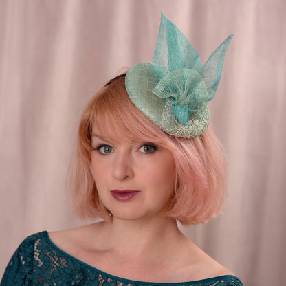 A woman with short pink hair wears a duck egg and teal horse racing fascinator hat with with lace and sinamay trim, photographed against a soft beige curtain background.