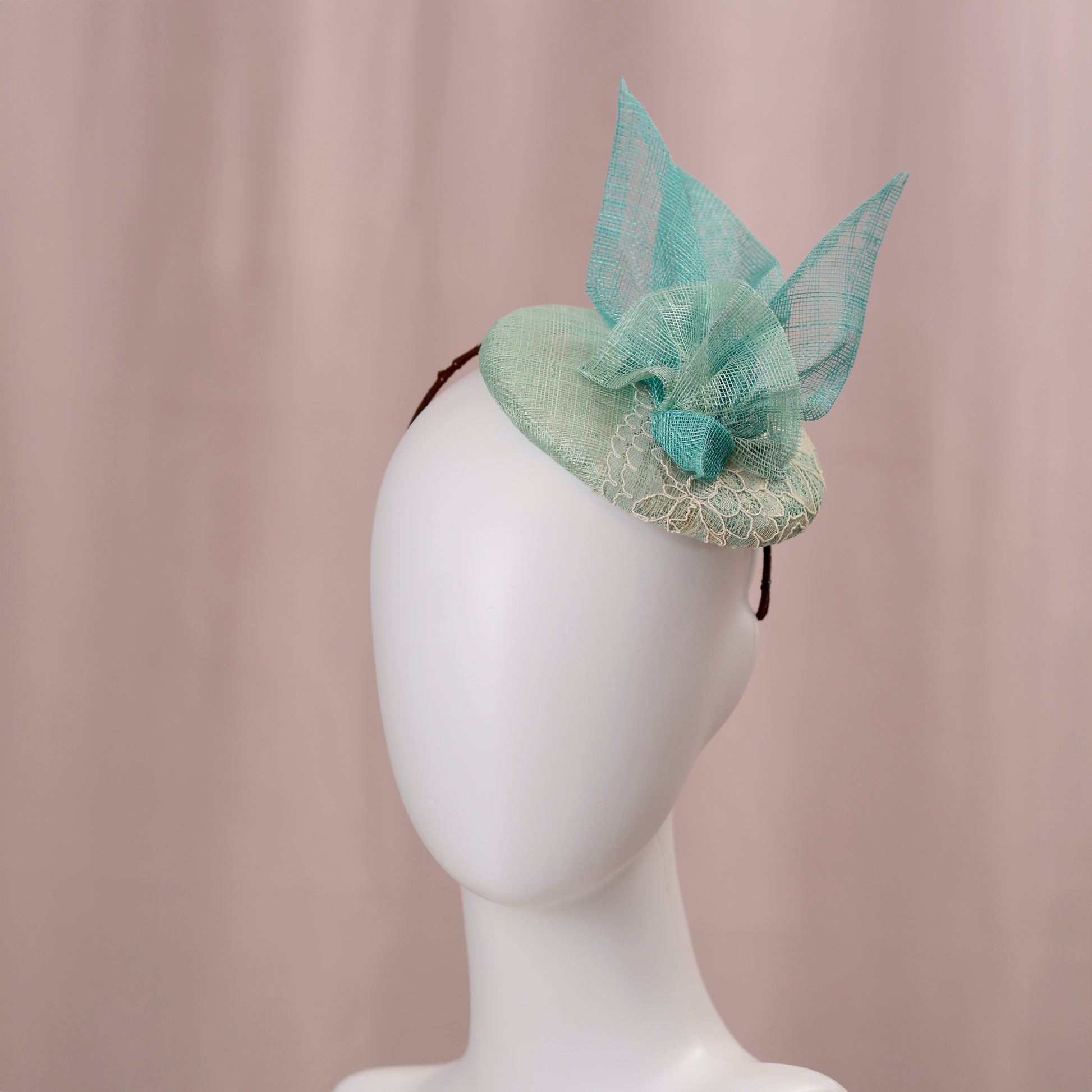 A mint green  and turquoise teal fascinator headpiece with sheer sinamay straw abstract bird wings and a lace trim, displayed on a white mannequin head against a soft pink background.