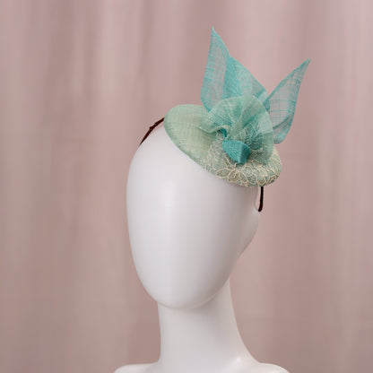 A mint green  and turquoise teal fascinator headpiece with sheer sinamay straw abstract bird wings and a lace trim, displayed on a white mannequin head against a soft pink background.