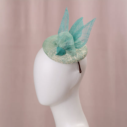 A mint green  and turquoise teal ladies day fascinator headpiece with sheer sinamay straw abstract bird wings and a lace trim, displayed on a white mannequin head against a soft pink background.