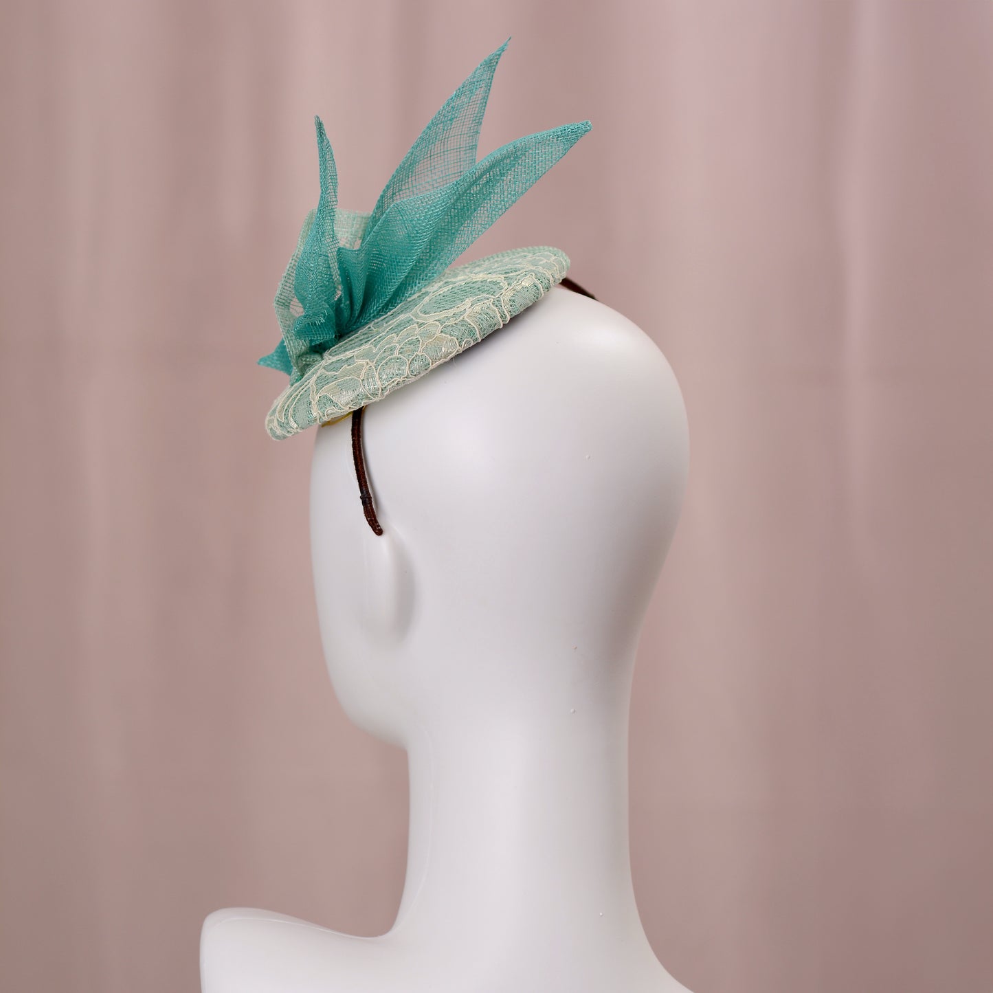 A mint green  and turquoise teal fascinator headpiece with sheer sinamay straw abstract bird wings and a lace trim, displayed on a white mannequin head against a soft pink background.