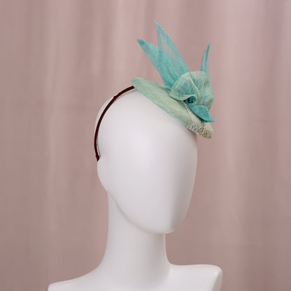 A mint green  and turquoise teal fascinator headpiece with sheer sinamay straw abstract bird wings and a lace trim, displayed on a white mannequin head against a soft pink background.