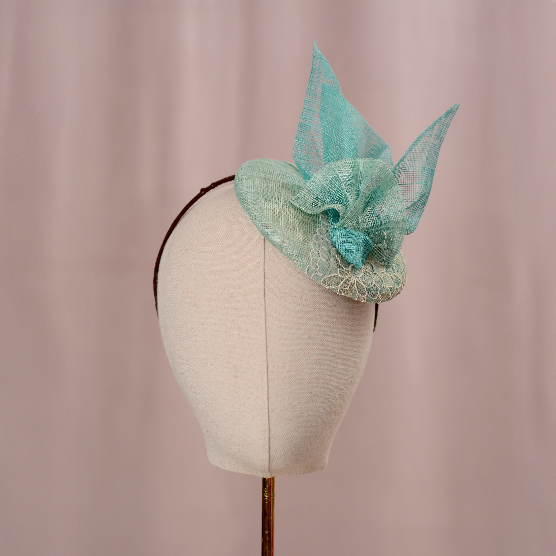 A mint green and turquoise teal royal ascot  fascinator headpiece with sheer sinamay straw abstract bird wings and a lace trim, displayed on a white mannequin head against a soft pink background.