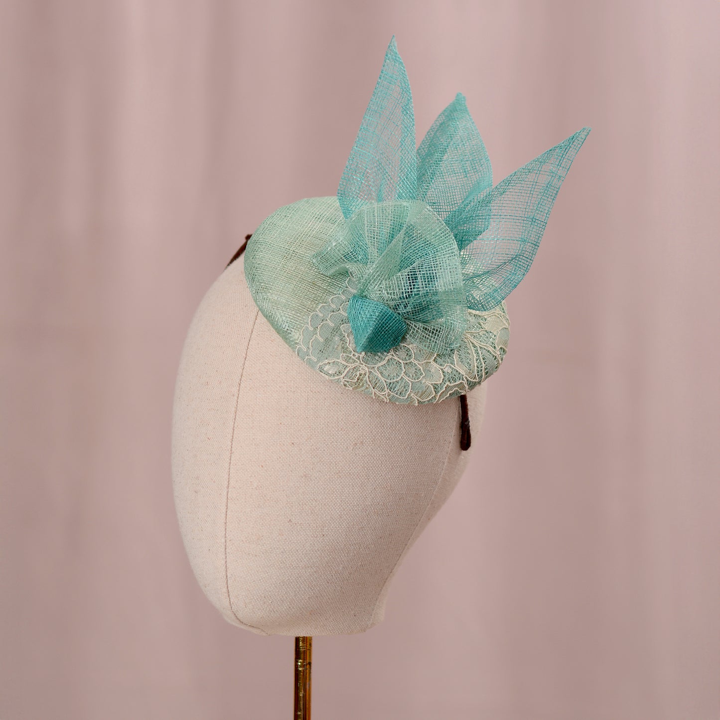 A mint green  and turquoise teal horse racing fascinator headpiece with sheer sinamay straw abstract bird wings and a lace trim, displayed on a white mannequin head against a soft pink background.