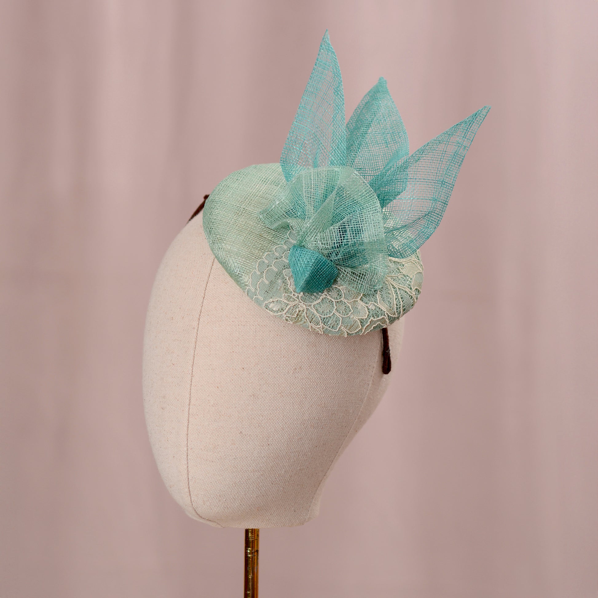 A mint green  and turquoise teal horse racing fascinator headpiece with sheer sinamay straw abstract bird wings and a lace trim, displayed on a white mannequin head against a soft pink background.