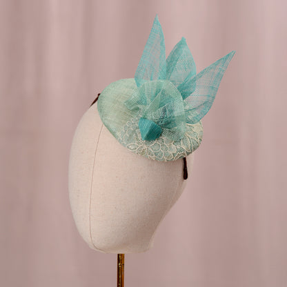 A mint green  and turquoise teal horse racing fascinator headpiece with sheer sinamay straw abstract bird wings and a lace trim, displayed on a white mannequin head against a soft pink background.