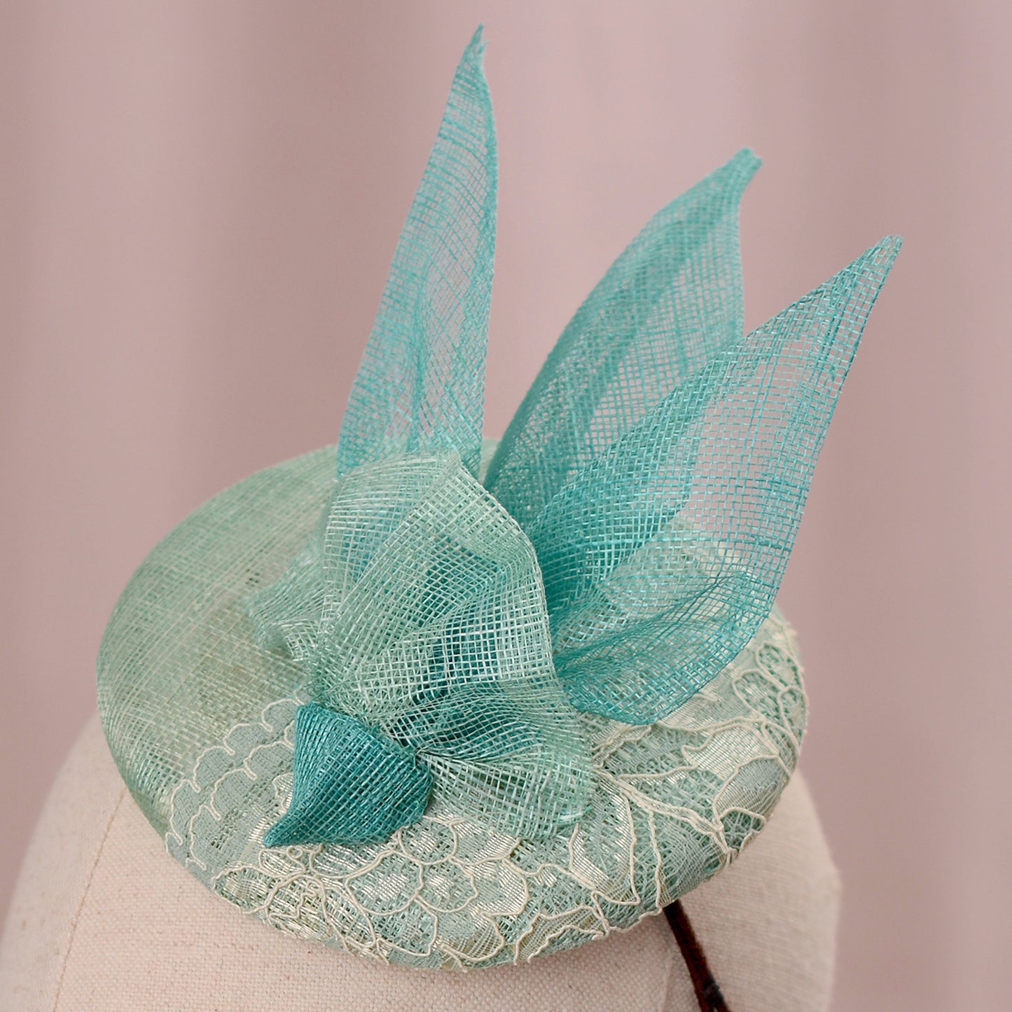A mint green  and turquoise teal fascinator headpiece with sheer sinamay straw abstract bird wings and a lace trim, displayed on a white mannequin head against a soft pink background.