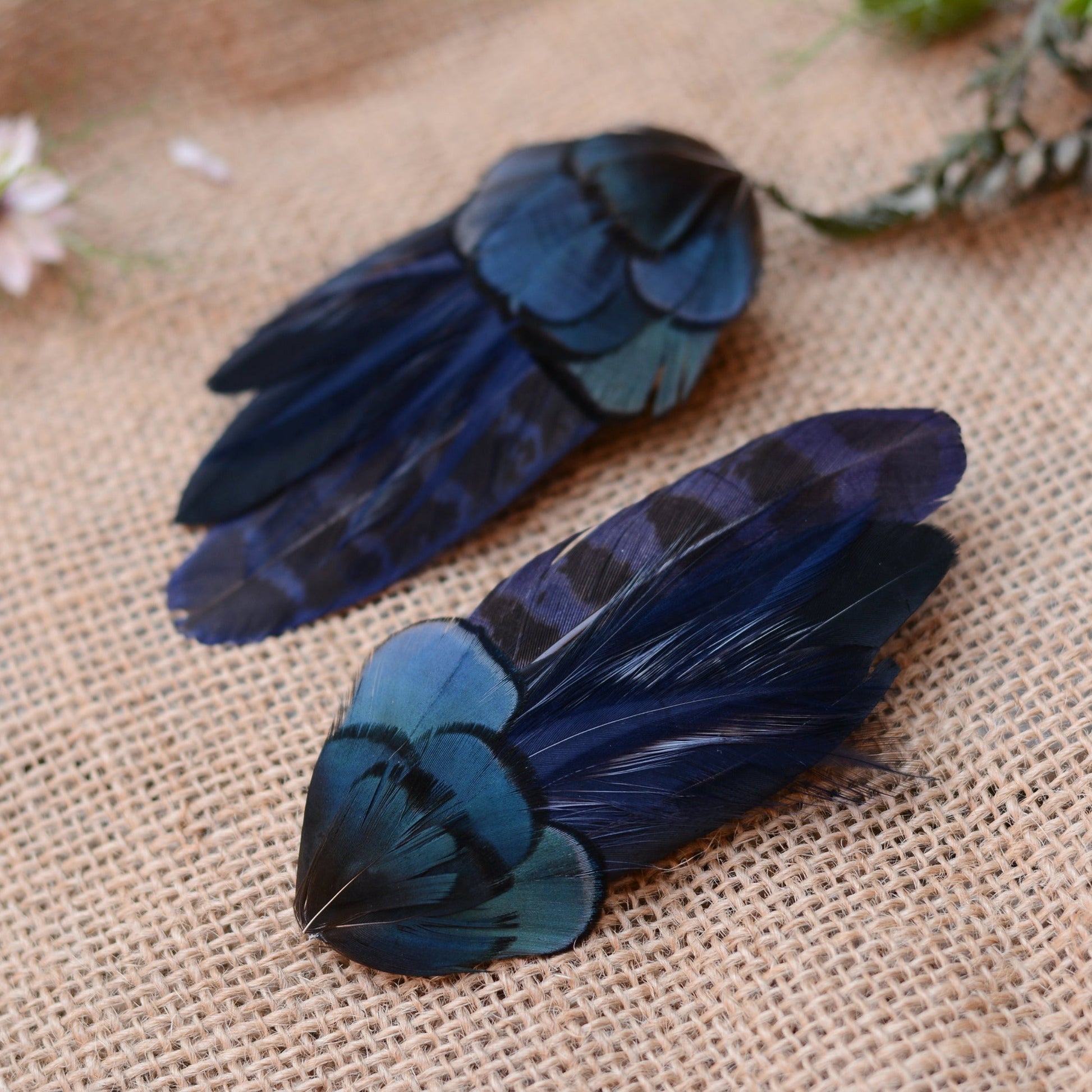Pair of hair clips crafted in a range of deep blue and black feathers showing different angles on a textured beige surface with flowers in the background