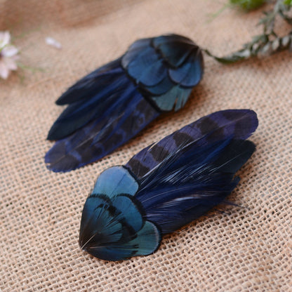 Pair of hair clips crafted in a range of deep blue and black feathers showing different angles on a textured beige surface with flowers in the background