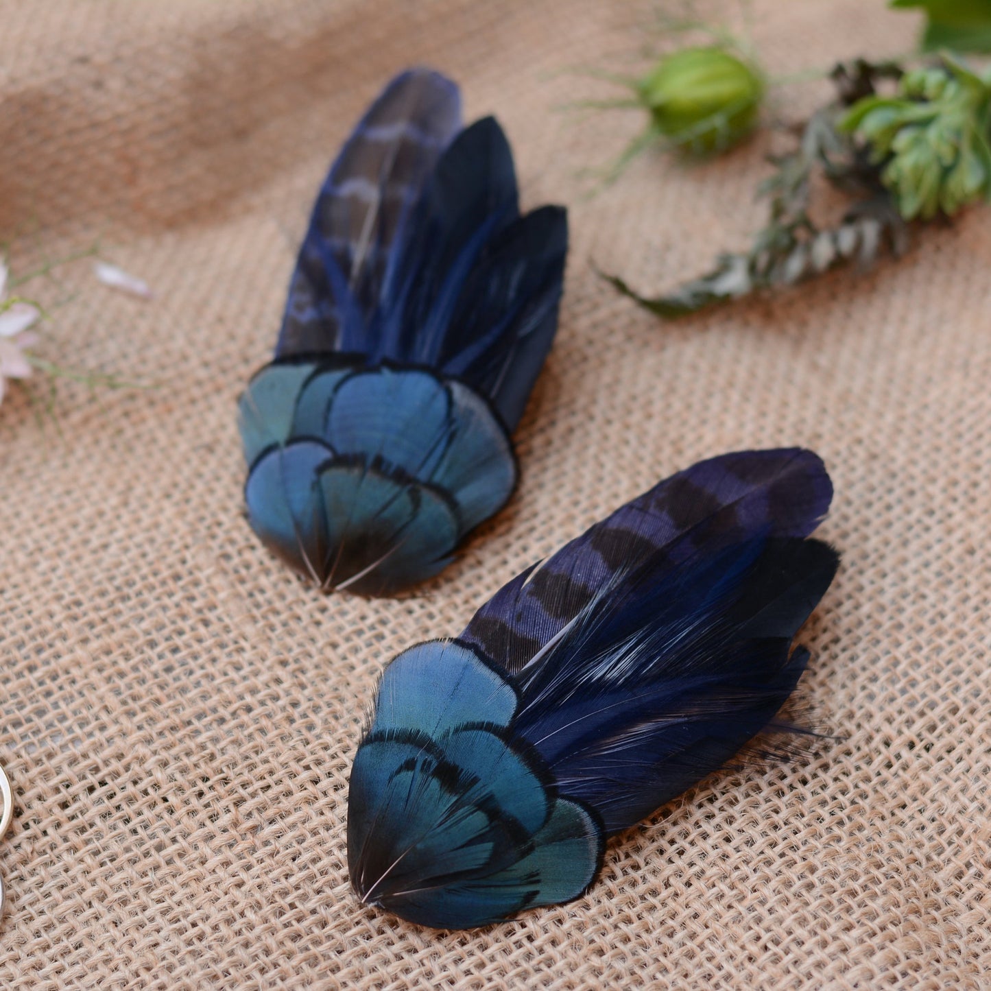 Pair of hair clips crafted in a range of deep blue and black feathers showing different angles on a textured beige surface with flowers in the background