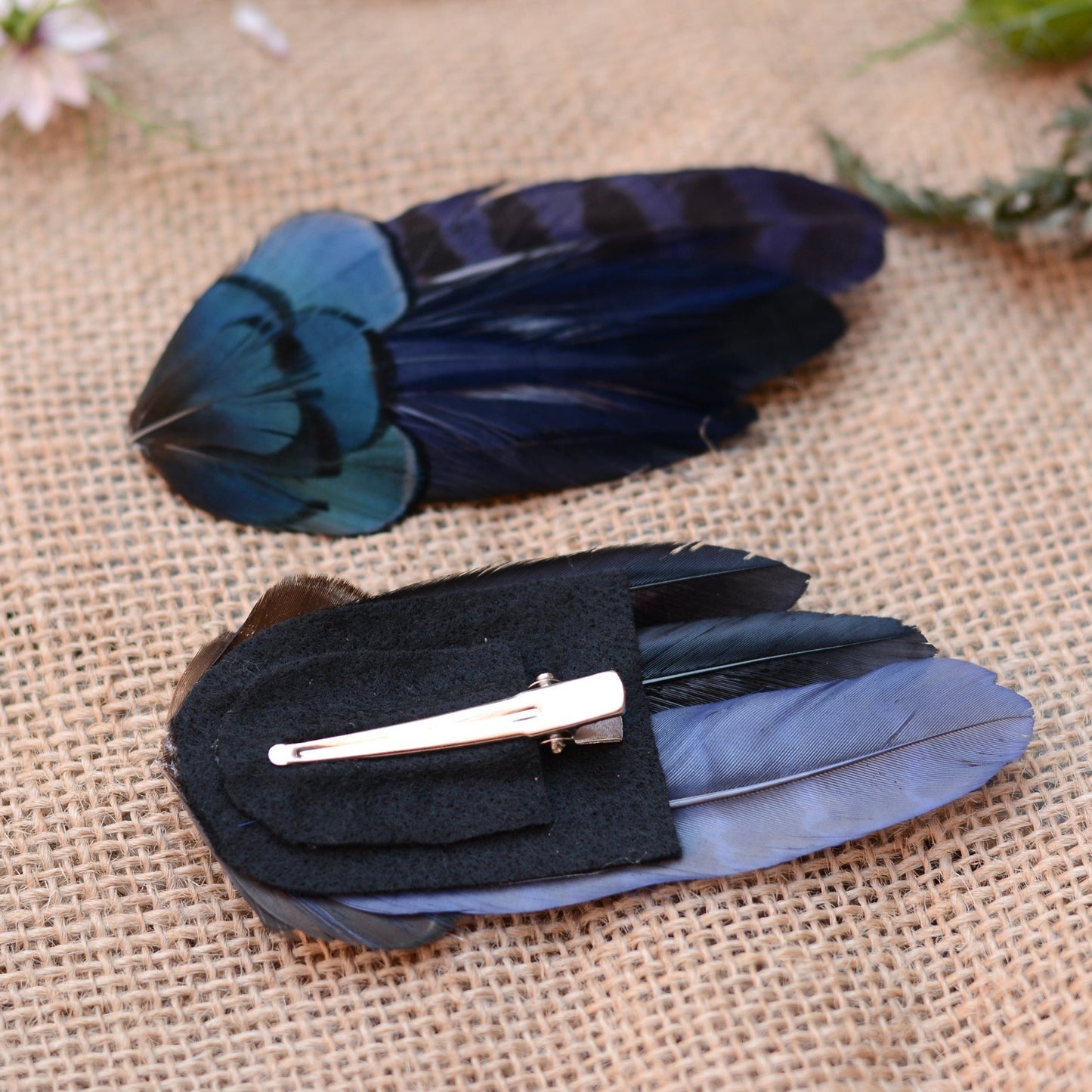 Pair of hair clips crafted in a range of deep blue and black feathers one is upside down to show the corcodile style clip attachment - laying on a textured beige surface with flowers in the background