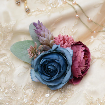 A decorative hair clip floral arrangement featuring a large blue rose, a deep burgundy ranunculus, pink chrysanthemums, and muted green leaves, styled on a cream lace bridal fabric background with a delicate necklace.
