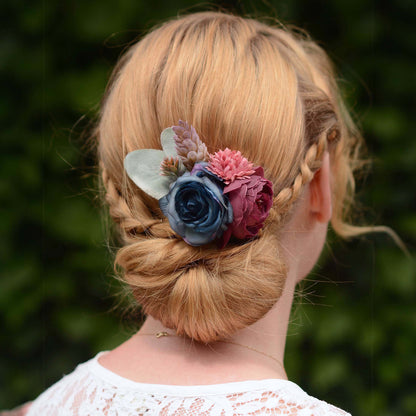 A blonde woman shown from the back wearing a braided low bun adorned with a decorative floral hair accessory featuring blue, pink, and burgundy faux flowers.