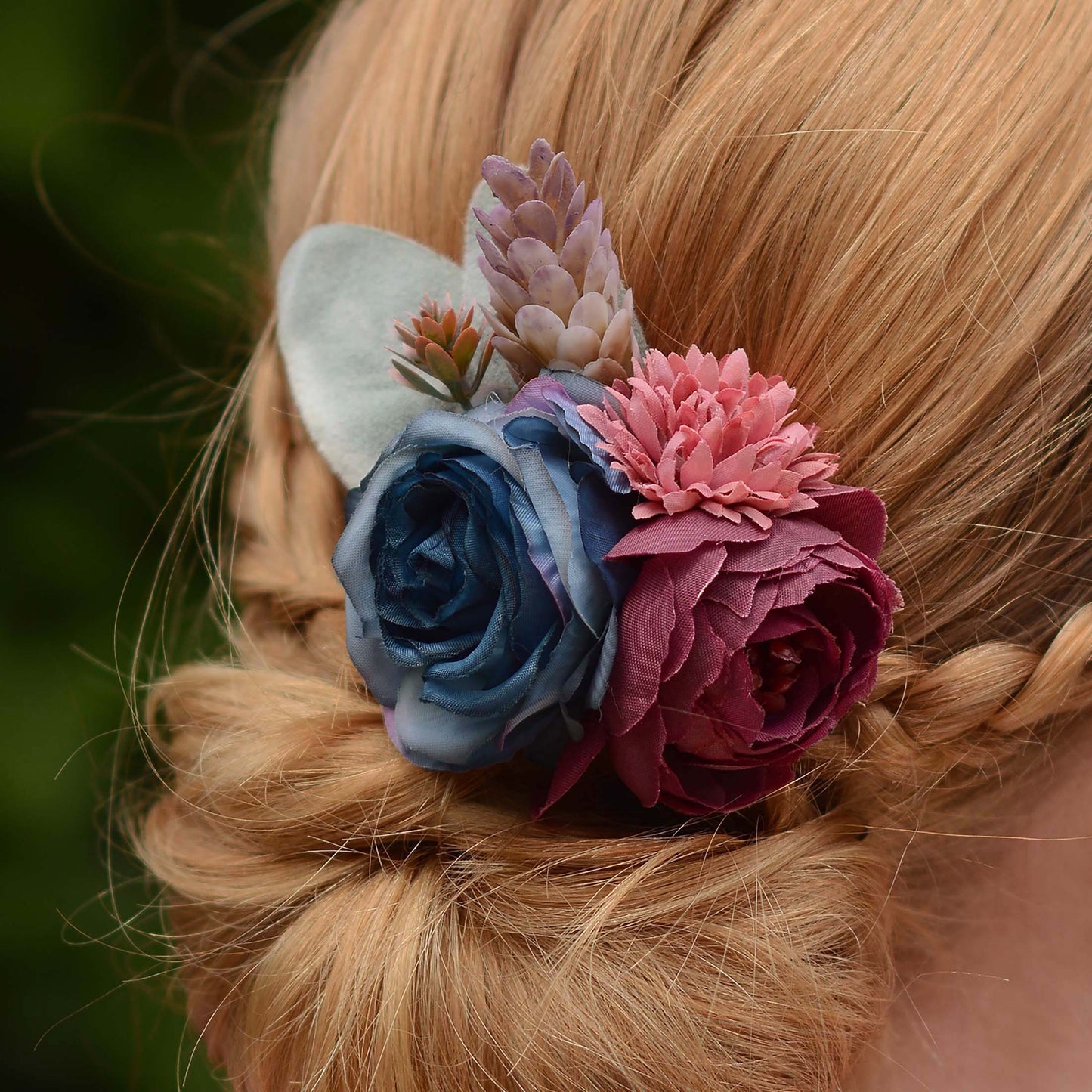 A close-up of a blonde braided hairstyle featuring a decorative hair accessory with layered fabric flowers in blue, burgundy, and dusty pink, plus a pale green leaf accent.