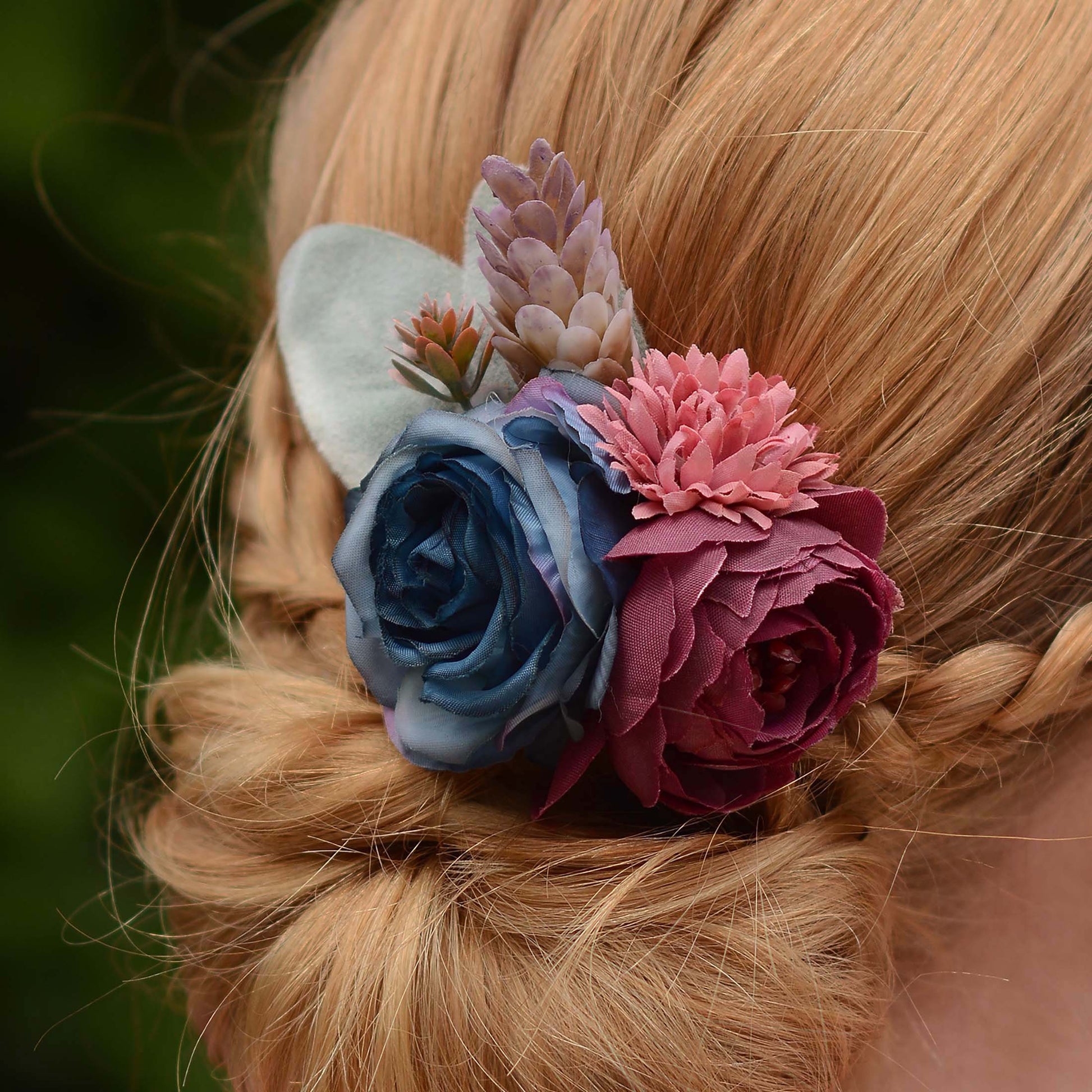A close-up of a blonde braided hairstyle featuring a decorative hair accessory with layered fabric flowers in blue, burgundy, and dusty pink, plus a pale green leaf accent.